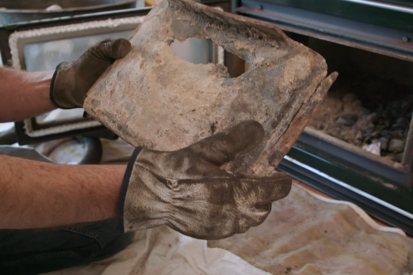 Gloved hands hold a worn wood burner baffle with a hole in it.