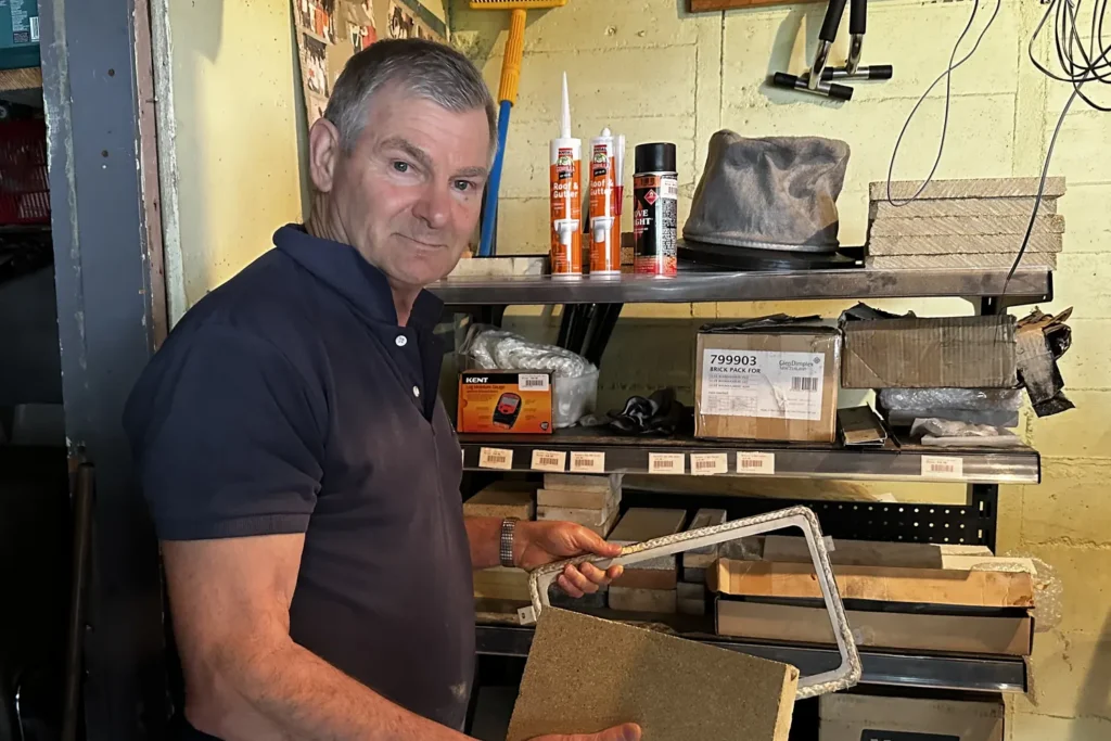 Alan from Chimney Magic holding a firebrick beside shelves of the wood burner parts he carries