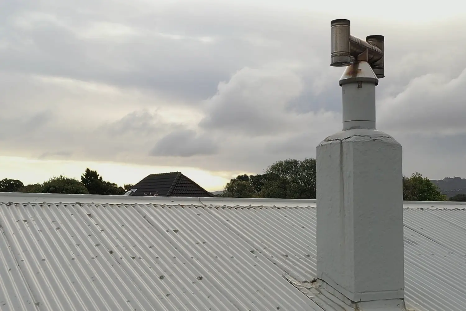 Wood burner flue cowl on a white-painted chimney above a corrugated iron roof in Lower Hutt